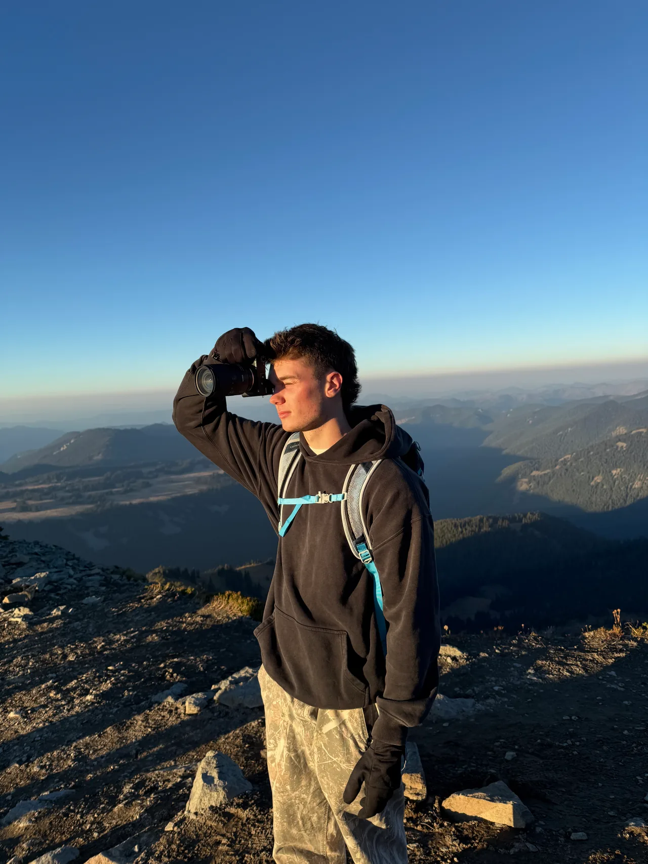Daniel Akselrod on a summit, shading his eyes, with a valley and ridgelines behind him.